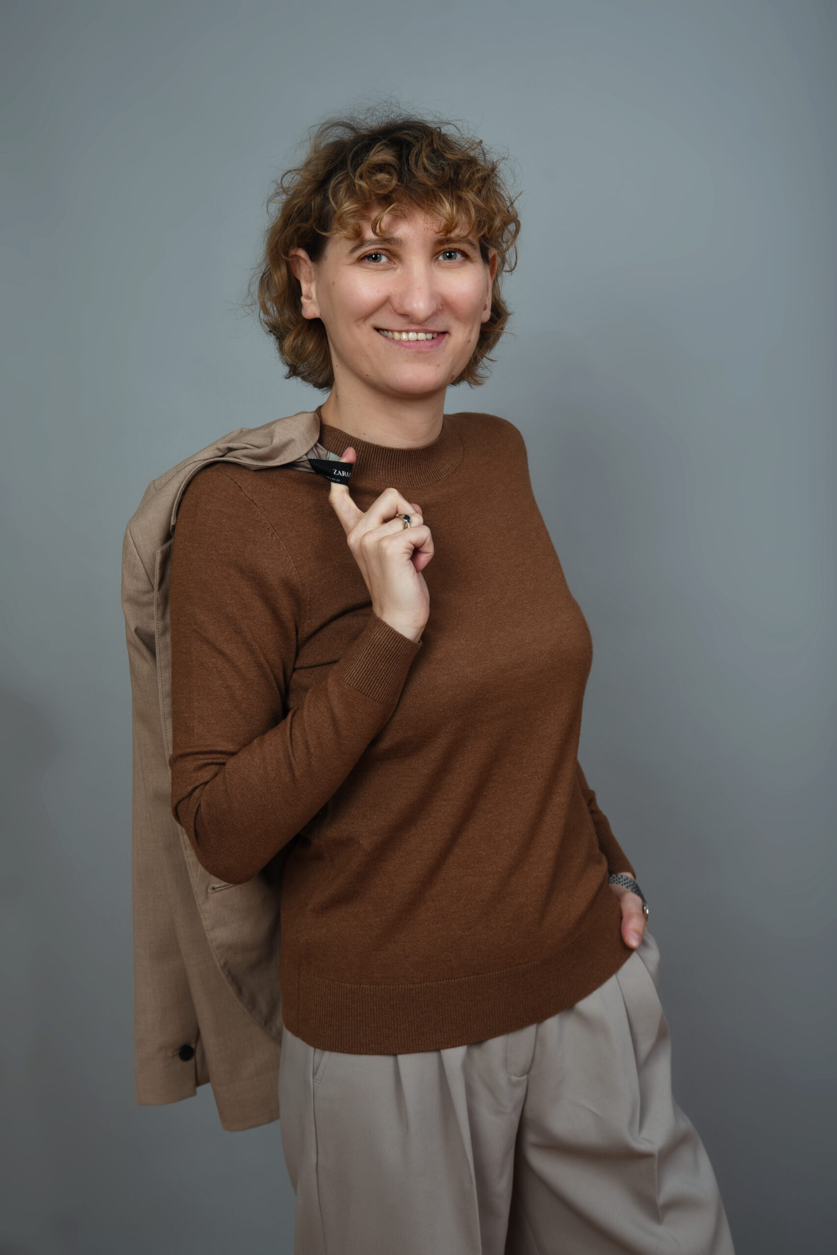 autorka bloga stoi w centrum zdjęcia, ma na sobie brązowy sweter i beżowe spodnie, uśmiecha sie i patrzy prosto w obiektyw, na ramieniu trzyma marynarkę