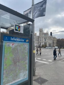 Na pierwszym planie wiata przystanku autobusowego z widocznym kodem NaviLens, w tle budynek banku w Madrycie