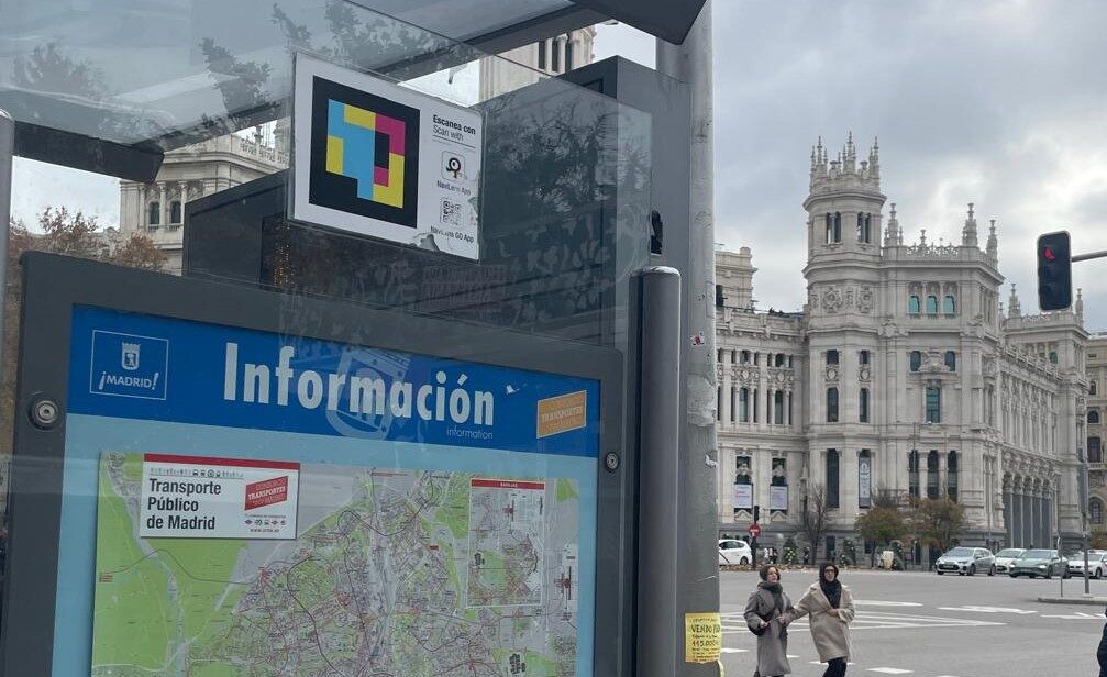 Na pierwszym planie przystanek autobusowy z widocznym kodem NaviLens - kolorowe kwadraty otoczone czarną obramówką, w tle budynek banku w Madrycie